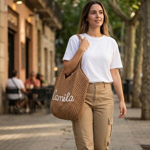 May include: A brown crocheted tote bag featuring the name Camila in white cursive script lettering. The bag has an open-weave texture and two shoulder straps, worn over the shoulder with a plain white t-shirt and tan cargo pants.