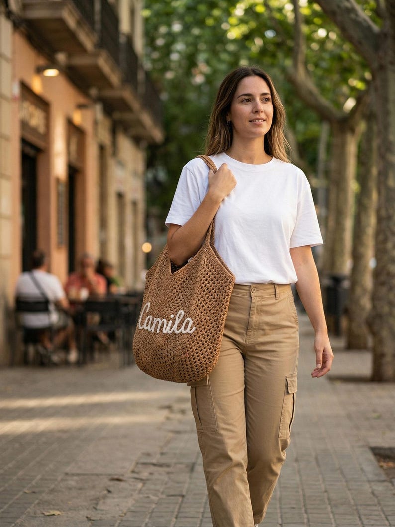 May include: A brown crocheted tote bag featuring the name Camila in white cursive script lettering. The bag has an open-weave texture and two shoulder straps, worn over the shoulder with a plain white t-shirt and tan cargo pants.