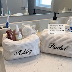May include: Two white waffle-textured fabric toiletry bags with black embroidered names. One bag features the name Ashley and contains a facial spray, cotton rounds, and swabs. The other bag features the name Rachel. A label lists Bag Color: White, Bag Size: Large, Name Color: Black, Font: F7.