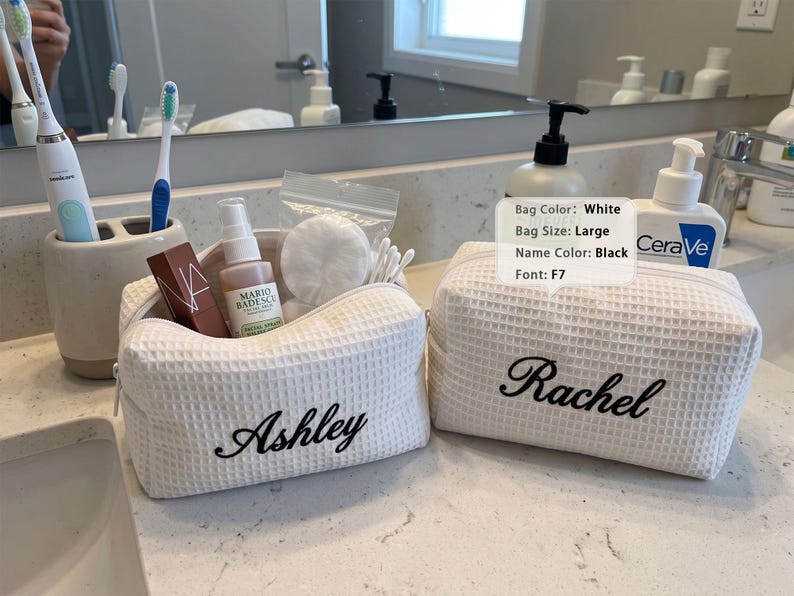 May include: Two white waffle-textured fabric toiletry bags with black embroidered names. One bag features the name Ashley and contains a facial spray, cotton rounds, and swabs. The other bag features the name Rachel. A label lists Bag Color: White, Bag Size: Large, Name Color: Black, Font: F7.