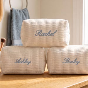 May include: Three rectangular cream-coloured waffle-textured toiletry bags with a zip closure. Each bag features a name embroidered in blue script font on the front: Rachel on the top bag, and Ashley and Bailey on the two bottom bags.