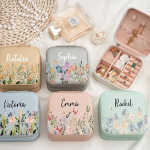 May include: A collection of personalized jewelry travel cases in various colors, including beige, gray, blue, and pink. Each case features a floral design and a name in script. One case is open, displaying jewelry.
