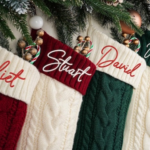 May include: Four cable-knit Christmas stockings in red, cream, and green, each with a name embroidered in a contrasting color: Juliet, Stuart, David, and William. Festive holiday decor with greenery and ornaments.