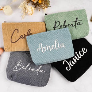 Puede incluir: Cinco neceseres de maquillaje de pana en varios colores: beige, verde, azul, gris y negro. Cada bolsa tiene un nombre bordado en un color contrastante. También se ven brochas de maquillaje y perfume.