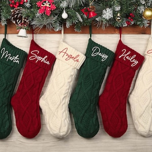 May include: Six cable-knit Christmas stockings in green, red, and cream, each with a name embroidered in a contrasting color. The stockings hang from a wooden ledge decorated with festive greenery and ornaments.