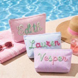 May include: Three rectangular zippered pouches made of seersucker fabric in pink and white, blue and white, and purple and white stripes. Each pouch features a personalised name embroidered in script lettering on the front: Edith, Lauren, and Vesper.