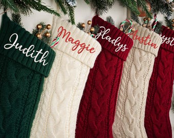 May include: Five cable-knit Christmas stockings in green, cream, and red, each with a name embroidered in a contrasting color. The stockings are decorated with candy canes and festive greenery.