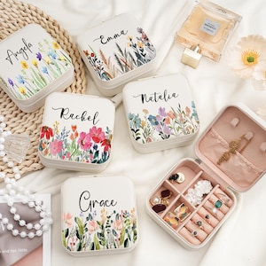 Puede incluir: Cajas de joyería cuadradas blancas con diseños florales y nombres personalizados como "Angela" y "Emma". Una caja está abierta, revelando anillos y pendientes. Las cajas están dispuestas sobre una superficie blanca con una estera tejida y un collar de perlas.