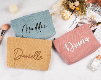 May include: Three corduroy cosmetic bags in blue, mustard yellow, and pink, each embroidered with a name in a cursive font. The bags are rectangular with a zipper closure. Makeup brushes and a perfume bottle are also visible.