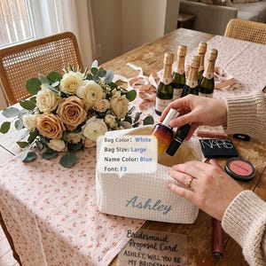 May include: A large white waffle-textured bag with the name Ashley embroidered in blue script font. The bag is open and contains a small bottle and a black lipstick tube. A clear acrylic card reads: Bridesmaid Proposal Card ASHLEY, WILL YOU BE MY BRIDESMAID?