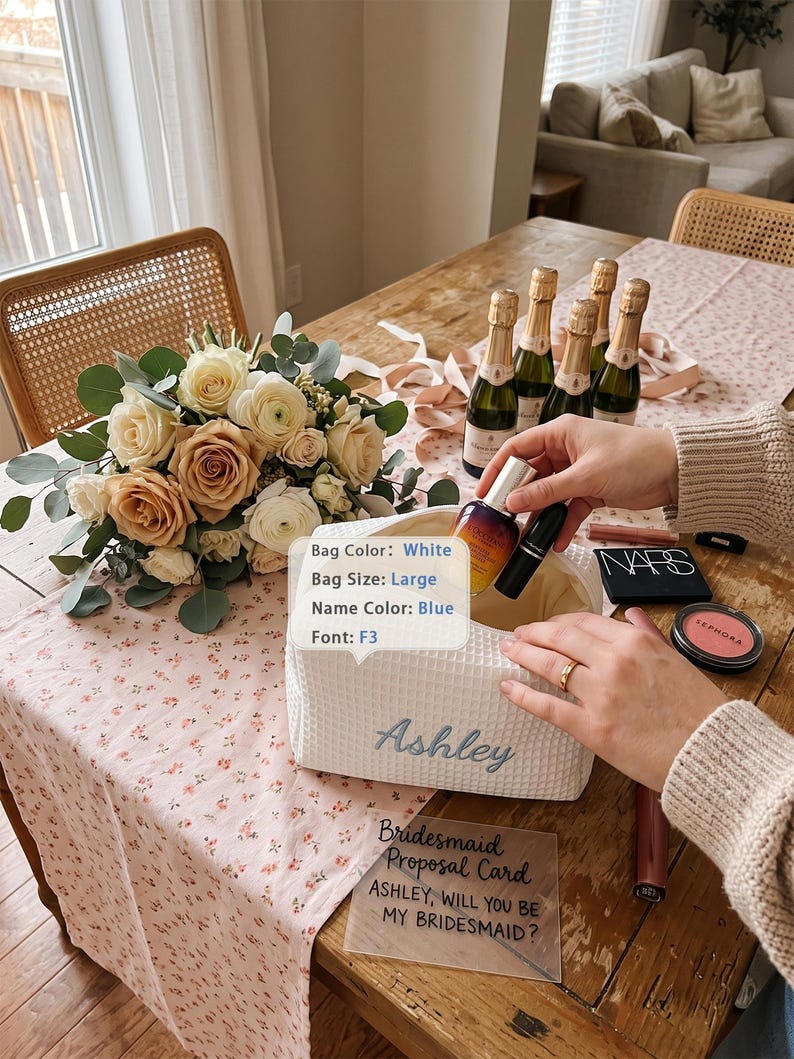 May include: A large white waffle-textured bag with the name Ashley embroidered in blue script font. The bag is open and contains a small bottle and a black lipstick tube. A clear acrylic card reads: Bridesmaid Proposal Card ASHLEY, WILL YOU BE MY BRIDESMAID?