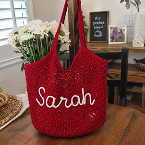 May include: A deep red crochet tote bag featuring an open-work mesh pattern and two shoulder straps. The front of the bag is embellished with the name Sarah written in white cursive script lettering made from a thick, textured yarn material.