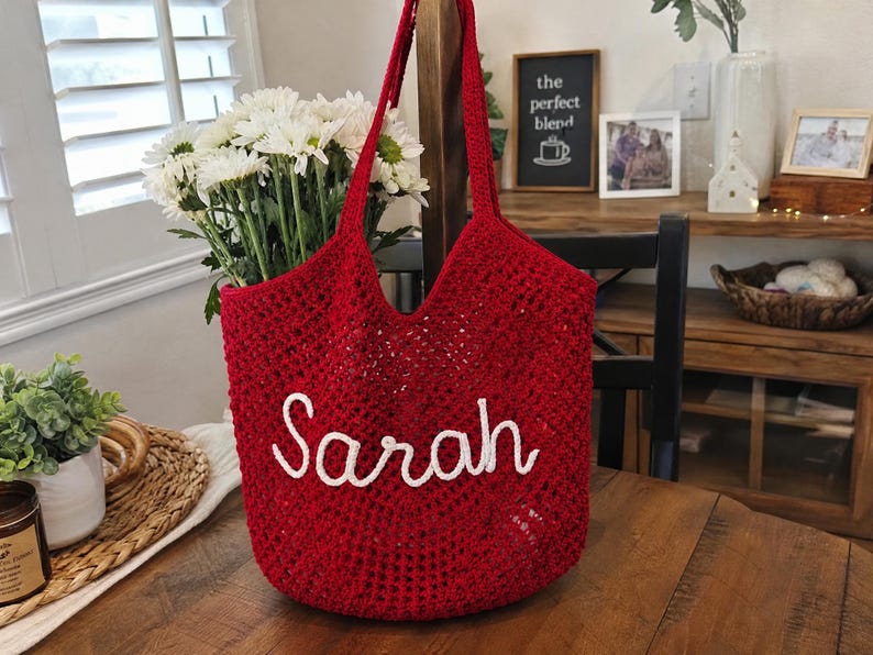 May include: A deep red crochet tote bag featuring an open-work mesh pattern and two shoulder straps. The front of the bag is embellished with the name Sarah written in white cursive script lettering made from a thick, textured yarn material.