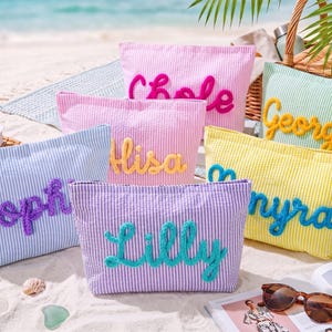 May include: A collection of seersucker cosmetic bags in various pastel colors, each embroidered with a different name in a contrasting color. The bags are arranged on a sandy beach, suggesting a summer theme.