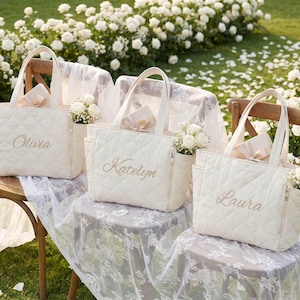Pode incluir: Três bolsas tote acolchoadas em tom off-white com bolsos laterais e bordados dourados. Cada bolsa é personalizada com um nome: Olivia, Katelyn ou Laura. As bolsas são decoradas com pequenos arranjos de flores brancas e fitas bege.