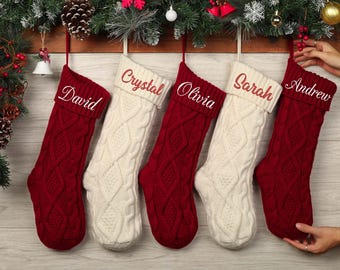 May include: Five cable-knit Christmas stockings in red and cream colors hang from a wooden beam decorated with greenery and ornaments. Each stocking is personalized with a name embroidered in a complementary color: David, Crystal, Olivia, Sarah, and Andrew.