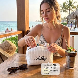May include: A large white waffle-textured cosmetic bag featuring the name Ashley embroidered in black script. The bag has a zipper closure and is positioned on a wooden table next to a pair of sunglasses and a drink in a metal cup.