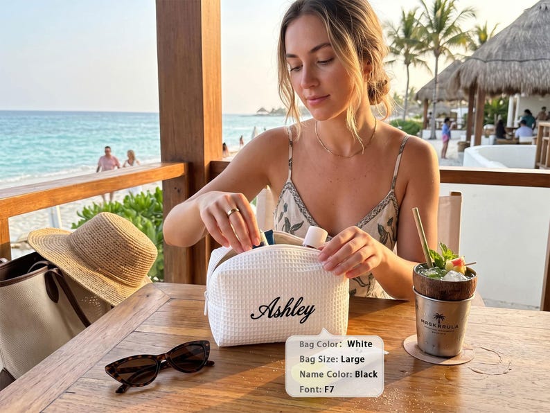 May include: A large white waffle-textured cosmetic bag featuring the name Ashley embroidered in black script. The bag has a zipper closure and is positioned on a wooden table next to a pair of sunglasses and a drink in a metal cup.