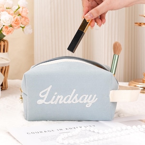 Puede incluir: Bolsa de cosméticos azul claro con el nombre "Lindsay" bordado en blanco. Una mano sostiene un labial negro y dorado sobre la cremallera abierta. Se ve un pincel de maquillaje.