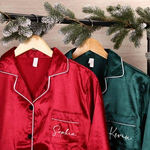 May include: Red and green satin pajama shirts hanging on a black rack, decorated with frosted pine garland. The red shirt has the name "Sophia" embroidered on the pocket. The green shirt has the name "Kevin" embroidered on the pocket.