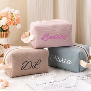 May include: Three personalised cosmetic bags in pink, taupe, and blue. Each bag has a zip closure, a handle, and a name embroidered on the front. The bags are arranged on a white surface with a pearl necklace.