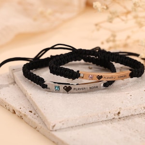 May include: Two matching black braided bracelets with engraved metal nameplates. One bracelet has a pink gem and the text "PLAYER 2 SAMUEL" with a heart symbol. The other has a blue gem and the text "PLAYER 1 NORA" with a heart symbol.