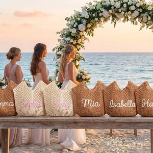May include: Six woven straw tote bags arranged in a row on a wooden table. Each bag features a unique name embroidered in white or pink script lettering, including the names Emma, Sophia, Ava, Mia, Isabella, and Harper, with an open-top design.