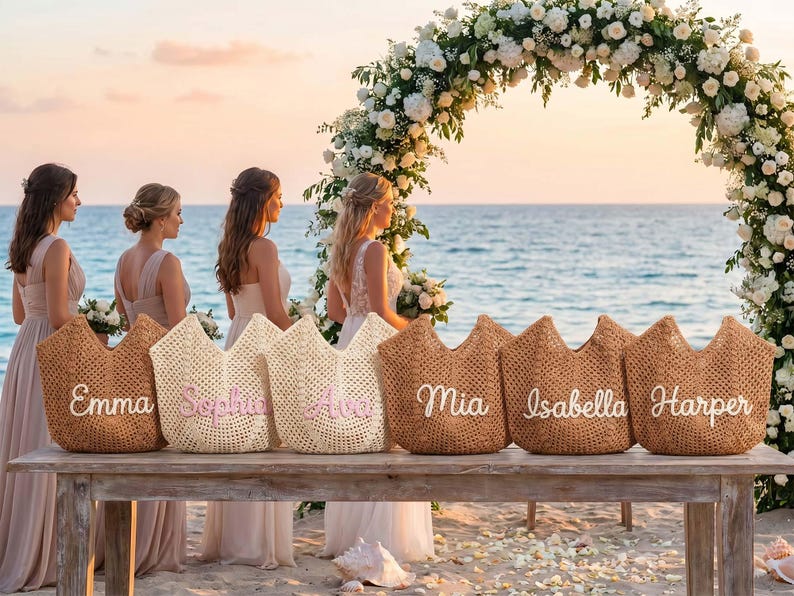 May include: Six woven straw tote bags arranged in a row on a wooden table. Each bag features a unique name embroidered in white or pink script lettering, including the names Emma, Sophia, Ava, Mia, Isabella, and Harper, with an open-top design.