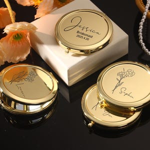 Op de afbeelding: Gouden compacte spiegels met gepersonaliseerde inscripties. Eén spiegel heeft de tekst "Jessica Bridesmaid 2025.4.30". Andere spiegels hebben bloemmotieven en namen. De spiegels liggen op een witte doos en een zwart oppervlak.