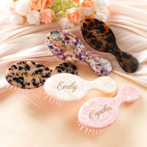 May include: Five personalized hairbrushes in various colors and patterns. The brushes are oval-shaped with names like "Bonnie," "Sophia," "Emily," "Cynthia," and "Adeline" in gold script. The background includes flowers and a satin fabric.