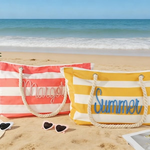 Op de afbeelding: Twee gestreepte strandtassen met touwhandvatten op een zandstrand. Eén is koraal en wit met "Margot" in cursief, de andere is geel en wit met "Summer" in cursief. Zonnebrillen en een hoed liggen in de buurt.