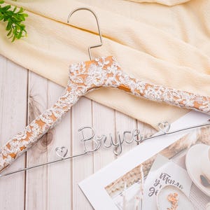 May include: A wooden clothes hanger with a silver hook and a decorative lace design. The hanger has the name "Bryce" in silver wire. The lace is white and covers the wooden portion of the hanger. The background includes a cream-colored fabric and a wooden surface.