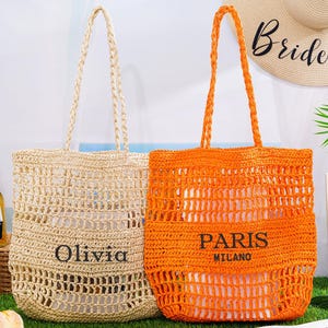 Op de afbeelding: Twee geweven draagtassen. Eén is beige met "Olivia" erop gedrukt, de andere is oranje met "Paris Milano" erop gedrukt. Beide hebben gevlochten handvatten. Op de achtergrond een strohoed met "Bride to be" erop gedrukt.