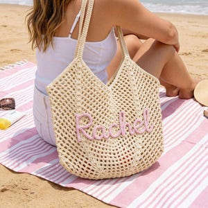May include: A beige crochet tote bag featuring an open-weave mesh design and two shoulder straps. The front of the bag is decorated with the name Rachel written in raised, light pink cursive yarn lettering, creating a textured appearance.
