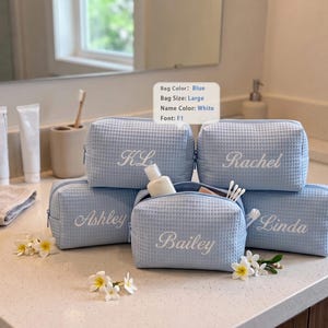 May include: Five light blue waffle-textured toiletry bags with white embroidered names including KL, Rachel, Ashley, Bailey, and Linda. A small sign displays Bag Color: Blue, Bag Size: Large, Name Color: White, Font: F1. The bags feature zipper closures.