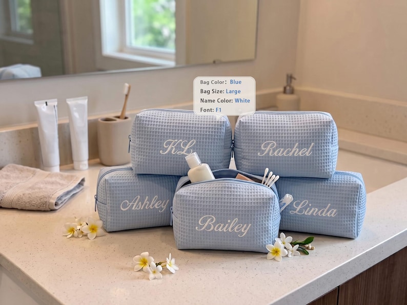 May include: Five light blue waffle-textured toiletry bags with white embroidered names including KL, Rachel, Ashley, Bailey, and Linda. A small sign displays Bag Color: Blue, Bag Size: Large, Name Color: White, Font: F1. The bags feature zipper closures.