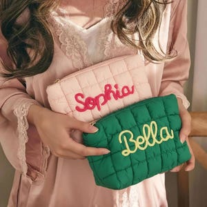 Op de afbeelding: Twee gewatteerde make-up tasjes. Eén is lichtroze met de naam "Sophia" in rood geborduurd. De andere is groen met "Bella" in geel geborduurd. Beide hebben gouden ritsen en een ruitpatroon.