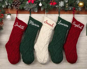 May include: Five cable-knit Christmas stockings in red, green, and cream colors hang from a wooden beam decorated with greenery and ornaments. Each stocking is personalized with a name in white script: Judith, Maria, Sarah, Lauren, and Crystal.