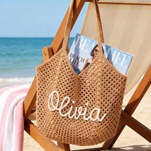 May include: A tan crochet tote bag featuring an open-weave design and two shoulder straps. The front of the bag displays the name Olivia in white, raised, cursive lettering. The bag is constructed from a textured, woven material.