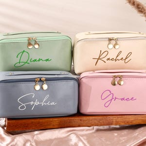 May include: Four personalised cosmetic bags in pastel shades: green, cream, blue, and pink. Each bag features a gold zip and a name embroidered in a matching colour: Diana, Rachel, Sophia, and Grace. The bags are displayed on a wooden surface.