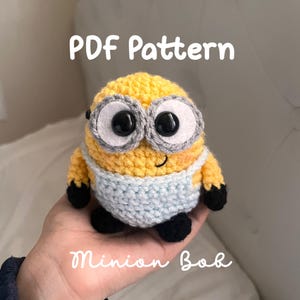 Op de afbeelding: Een gehaakte Minion Bob pop met een gele body, zwarte voeten en een lichtblauwe slabbetje. De pop heeft grote, ronde ogen met zwarte pupillen en grijze randen. De tekst "PDF Pattern" en "Minion Bob" is zichtbaar.