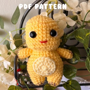 May include: A handmade, yellow crocheted dinosaur with black eyes and a red smile. The dinosaur has a cream-colored belly and is surrounded by greenery and white flowers. The text "PDF PATTERN" is visible at the top.