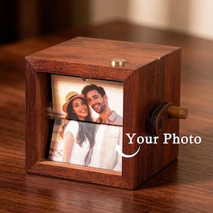 Puede incluir: Un cubo de fotos de madera marrón oscuro con un mecanismo giratorio, que muestra una foto de una pareja. El cubo tiene un pequeño botón de color latón en la parte superior. El texto "Your Photo" es visible.