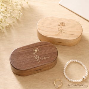 May include: Two wooden oval boxes, one light and one dark, with floral engravings and names. A pearl bracelet and a gold ring with a clear stone are also visible. The boxes are on a light wooden surface.