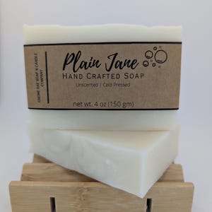 以下が含まれることがあります： 「Plain Jane」とラベルされた、無香料でコールドプレスされた石鹸が2つ、木製の石鹸置きに置かれています。石鹸はクリーム色で、ラベルには正味重量150gと記載されています。