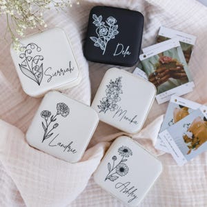May include: Four square jewelry cases in white and black, each featuring a unique black line-art floral illustration and a personalized name in script font. The names include Surriah, Dela, Monika, Landrie, and Ashley, with floral designs.