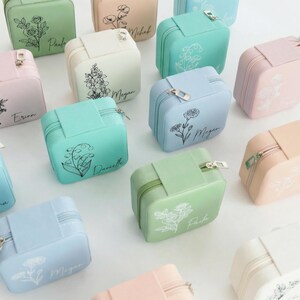 May include: A collection of square, zippered jewelry cases in pastel colors including mint, light blue, pale pink, and cream. Each case features a unique floral line art illustration and a personalized name printed in a cursive script font.