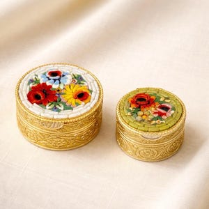 May include: Two small, round, decorative boxes with floral mosaic tops. The larger box features red, yellow, and blue flowers. The smaller box has red and yellow flowers on a green background. Both have gold-colored, ornate metal bases.