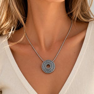 Puede incluir: Un collar con colgante circular de tono plateado que presenta un intrincado patr&oacute;n de volutas grabadas con un recorte central. El colgante cuelga de una cadena fina y lisa de tono plateado, sobre una prenda de tela acanalada clara.
