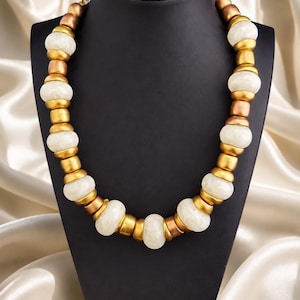 Puede incluir: Un collar llamativo con grandes cuentas ovaladas de color blanco roto, intercaladas con elementos metálicos dorados y cobrizos. El collar se exhibe sobre un maniquí negro, con un fondo de tela marfil drapeada.